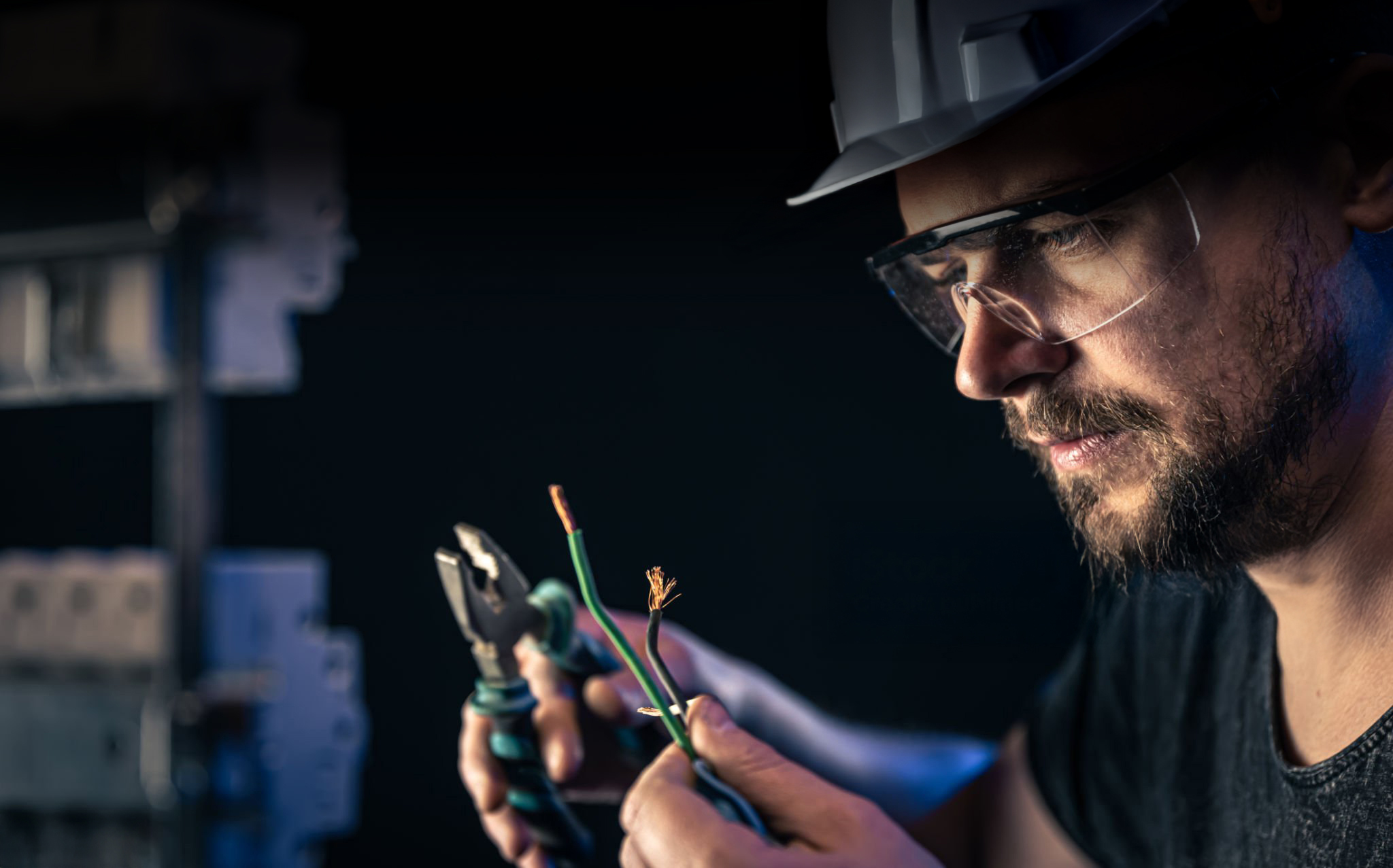 A male electrician in a protective helmet works in a switchboard with an electrical connecting cable.